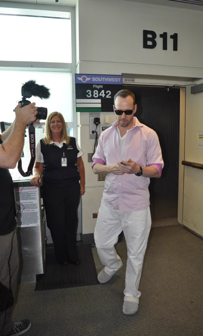 A person in sunglasses and casual clothing walking while looking at a phone at a Southwest Airlines gate labeled B11 for Flight 3842. A photographer is capturing the moment, and an airline staff member is smiling nearby.
