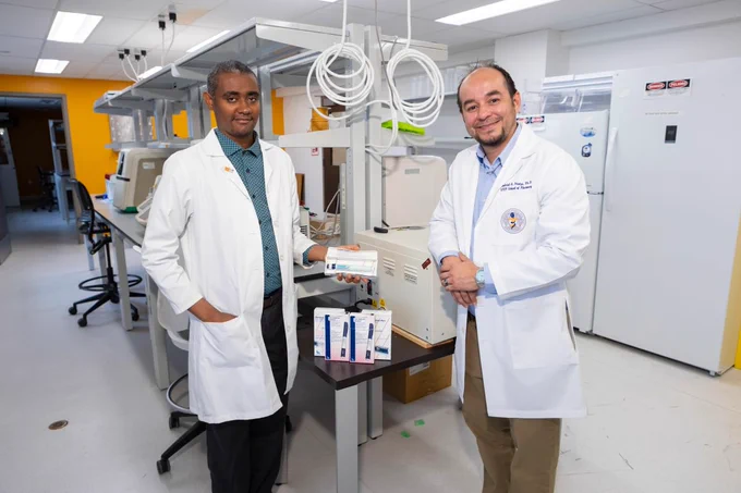 Two individuals in lab coats stand in a laboratory. One person is holding a prescription product, and on the table in front of them are several boxed items. Laboratory equipment and workspaces are visible in the background.
