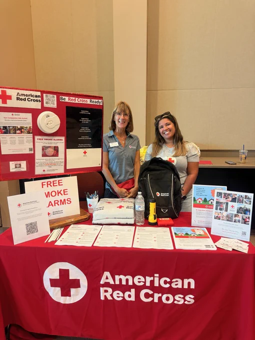 Two individuals smiling behind an American Red Cross booth featuring informational materials, a backpack, a flashlight, and a pillow. A sign reads "FREE SMOKE ALARMS." The table and backdrop display the American Red Cross logo and promotional posters.