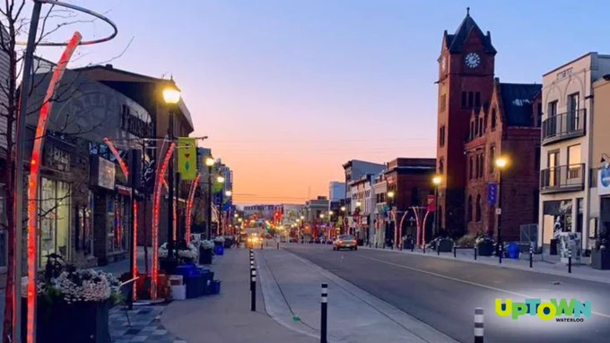 A street scene at dusk with the road lined with shops and decorated with illuminated street lights. Uptown Waterloo logo is displayed.
