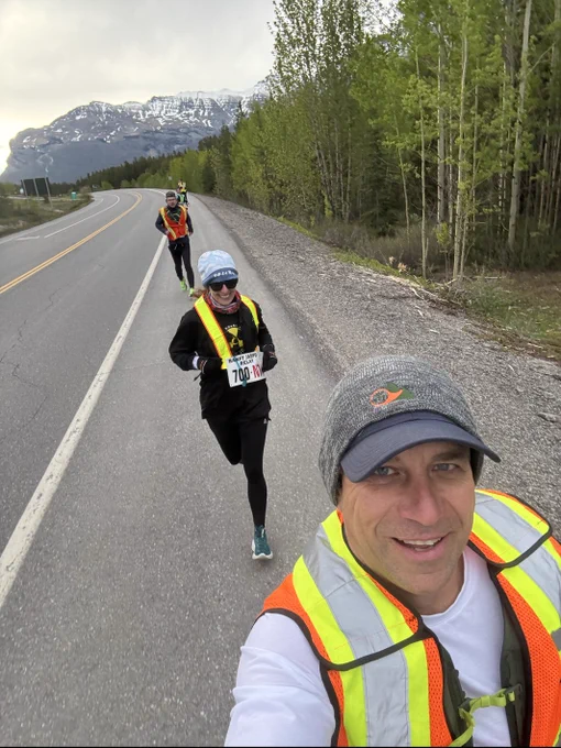 Runner wearing a race bib and reflective vest jogging on a roadside path with mountains and trees in the background.