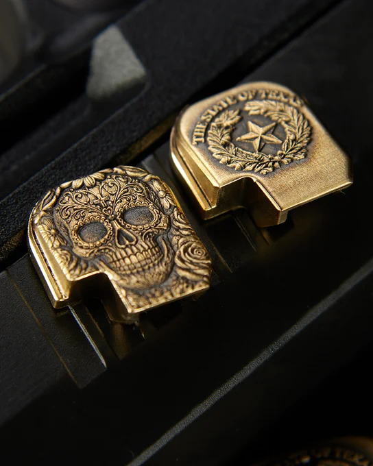 Two engraved brass pistol backplates displayed side by side on a black firearm slide. One features an ornate Sugar Skull design with floral artwork, while the other features the Texas state seal with a lone star and wreath engraving.