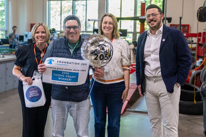 A group of four people celebrates a Thiems Grant win, holding signs and a balloon in a workshop setting. The mood is joyful and celebratory.