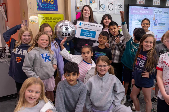 A joyful classroom scene with children and a teacher celebrating the 2026 Thiems Grant win. They are gathered around holding signs and balloons with smiles and excitement.