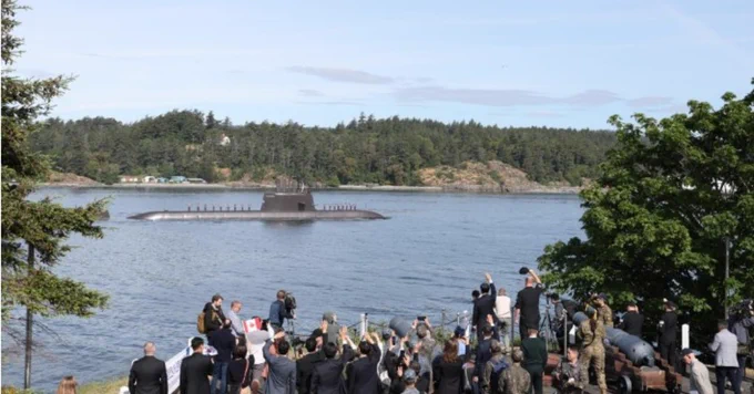 Crowd gathered on shore watching a submarine surfacing in calm waters near a forested coastline.