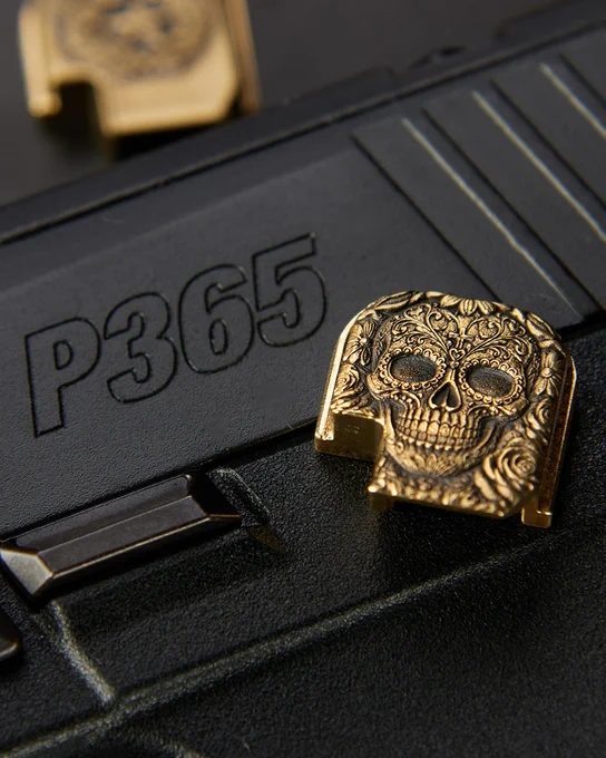 Close-up of a brass Sugar Skull engraved backplate installed on the rear of a black pistol slide. The detailed engraving features floral patterns, roses, and intricate decorative elements surrounding a stylized skull.