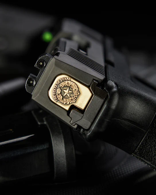 Brass Texas state seal backplate installed on the rear of a black pistol slide. The engraving features a lone star surrounded by a wreath with “The State of Texas” engraved along the top edge.