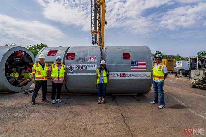 Four construction workers in safety gear stand beside a large Midwest Mole tunneling machine on a paved site under a partly cloudy sky. /