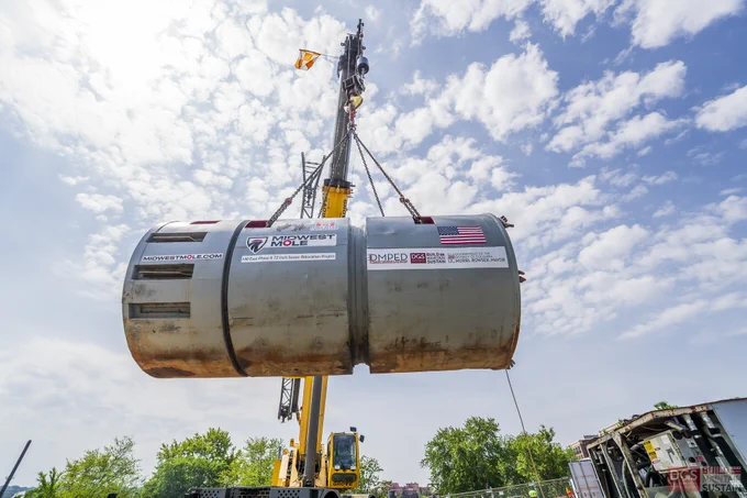 Large cylindrical tunnel boring machine being lowered into a deep circular excavation site surrounded by construction workers and equipment.