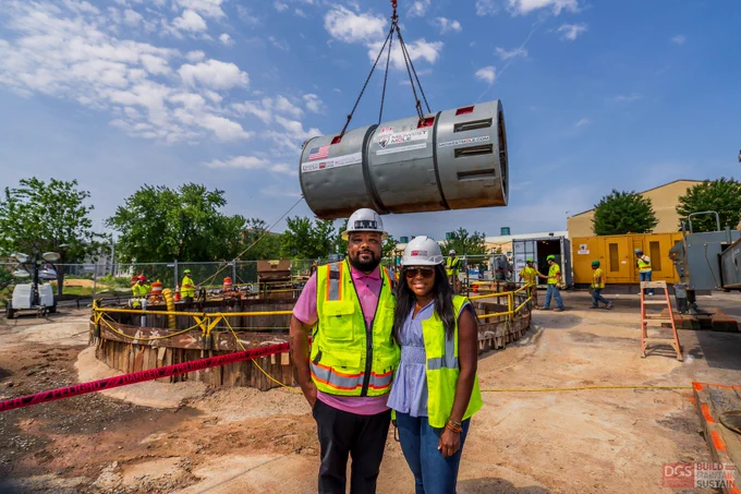 Two construction workers wearing safety vests and helmets stand in front of a large cylindrical metal structure being lifted by a crane at a busy construction site