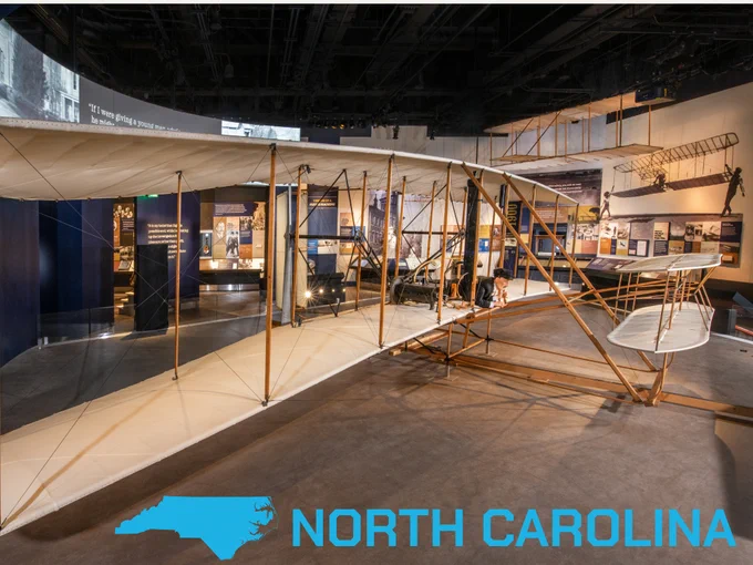 Historic biplane display at a museum with detailed exhibits in the background. The foreground features North Carolina state outline and the text "North Carolina".