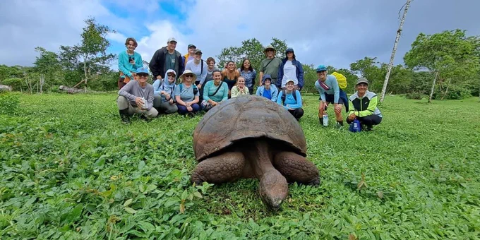 A group of SPC study abroad students in outdoor clothing sits on grass near a large tortoise in a lush green landscape at the Galápagos Islands.
