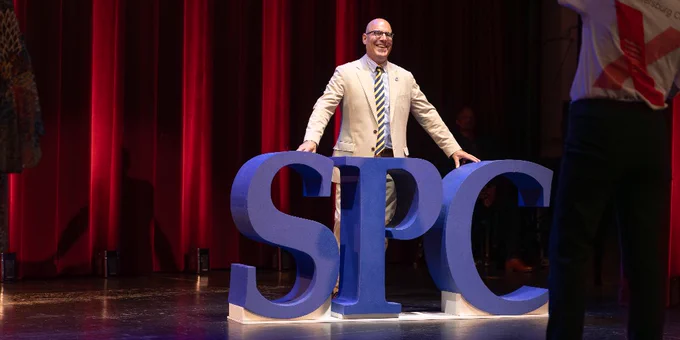 Jesse Turtle stands smiling behind large blue letters "SPC" on a stage with red curtains. Another individual is taking a photo from the side, wearing a TEDx T-shirt.