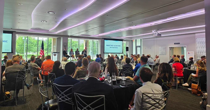 Conference room with audience seated at round tables as a panel discussion between 4 people on stage takes place.