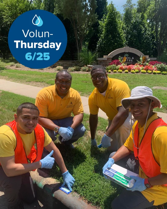 Four individuals in yellow shirts and safety vests are kneeling on grass beside a curb, participating in a community activity. A graphic at the top left corner reads "Volun-Thursday 6/25.