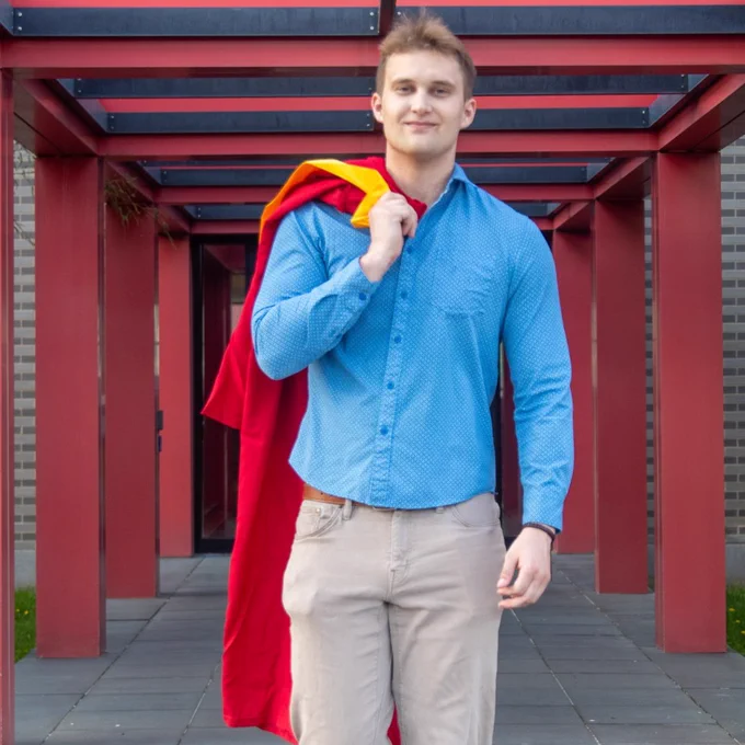 A person in a blue button-up shirt and beige pants is walking confidently under a modern, red-structured canopy. The individual is holding a red and yellow fabric over one shoulder. The surrounding area has brick walls and a tiled walkway.