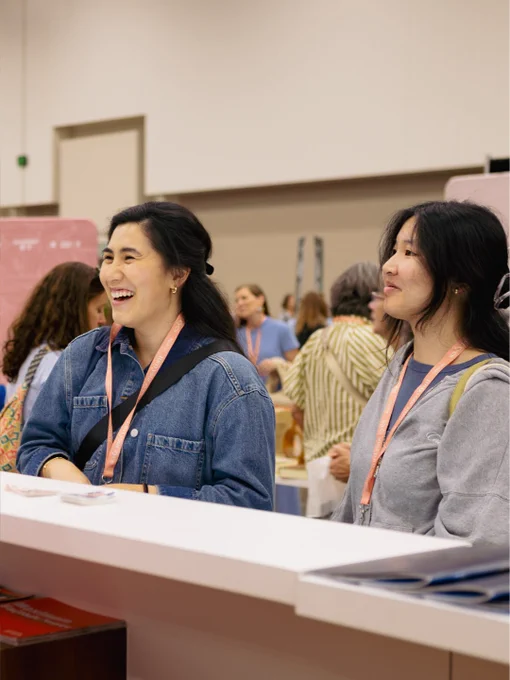Two women are seen smiling and conversing at a busy convention. They're wearing casual attire and lanyards indicating they are attendees.