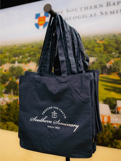 A display of Southern Seminary tote bags featuring the institution's logo and slogan. The bags hang on a rack with a backdrop showing a scenic view.