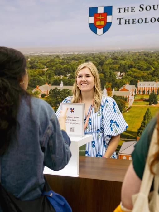 A woman interacts with attendees at a booth from a theological institute, engaging them with a giveaway opportunity.