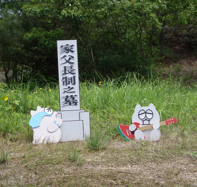草の生い茂る野外のちょっとした空間に、漢字で「かふちょうせいのはか」と縦書きで書かれた墓石のイラストボードと、横で泣き崩れるパワーストーンのブレスレットを手首に巻いた茶色いネコのイラストボードが立てられている。その50センチほど離れた場所に、我関せずといった感じでスイカ柄のエレキギターを夢中でかき鳴らす白いネコのイラストボードが立てられている。