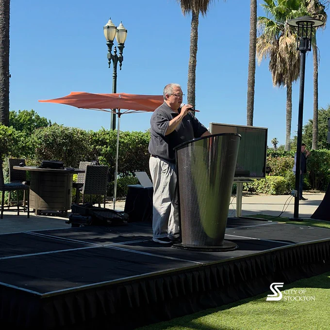 A person is speaking at an outdoor event, standing on a stage with a podium. The setting includes palm trees, chairs, tables, and a large umbrella. The logo "City of Stockton" is visible in the corner.