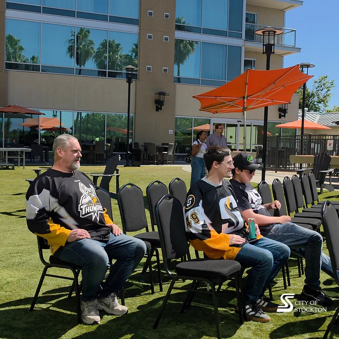 A group of people sitting on outdoor chairs in front of a modern building. Some are wearing hockey jerseys, one with "Thunder" on it. The setting is sunny with orange umbrellas and glass windows visible. The City of Stockton logo is in the bottom right corner.