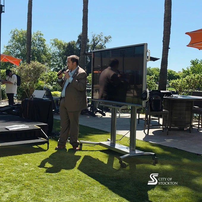 A person in a suit stands speaking into a microphone in an outdoor setting with greenery and orange umbrellas. They are near a large monitor on a stand. The backdrop features lush trees and a clear blue sky. A logo on the grass reads, "City of Stockton.