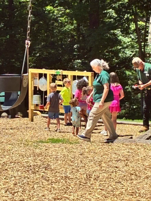 A group of children and two adults are at a playground. The children are interacting with a wooden activity wall. The area is surrounded by trees and wood chips cover the ground.