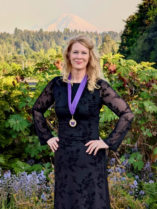 Blonde woman in a black floral dress with a purple ribbon and gold medal featuring a 'W' and '2023'. She stands with hands on hips, smiling, against a backdrop of lush green foliage and a distant snow-capped mountain under a bright sky.