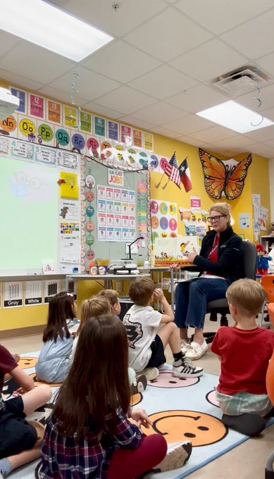 A female teacher with glasses reads from a book to a group of young children sitting on a smiley-face rug in a vibrant classroom. Yellow walls are adorned with alphabet charts.