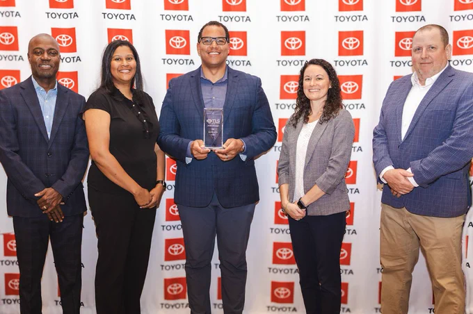 A group of five people standing in front of a backdrop featuring the Toyota logo. The person in the center is holding an award. All are dressed in business attire and smiling for the photo.