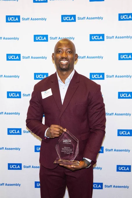 A person dressed in a burgundy suit and wearing a name tag holds an award. The backdrop features a branded step-and-repeat banner with "UCLA Staff Assembly" logos.