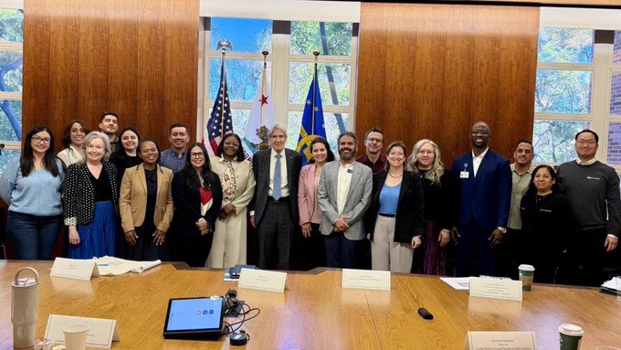 A group of individuals stands around a large wooden table in a professional meeting room. Flags of the United States, California, and another organization are visible near the windows at the back of the room. The group consists of people dressed in business and formal attire. Documents and coffee cups are placed on the table.