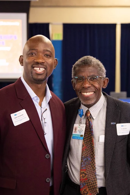 Two individuals pose together at an awards event. One person is wearing a burgundy suit, while the other is dressed in a patterned tie and formal attire. Both have name badges visible. The background includes blue curtains and a presentation screen.