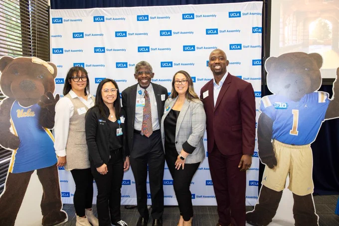 A group of individuals poses in front of a branded "UCLA Staff Assembly" step-and-repeat backdrop. Two large cardboard cutouts of bear mascots wearing UCLA apparel flank the group on both sides. The individuals are dressed in business and professional attire. A presentation screen is visible to the right.