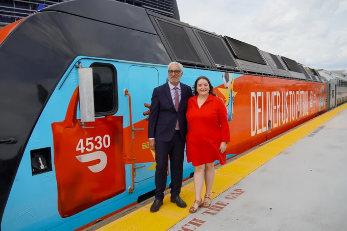 A person in formal attire and another in a red dress stand smiling in front of a NJ TRANSIT train at a stadium. The train's exterior is brightly colored with the words "Deliver Us to Futbol".