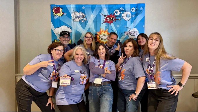 A group of nine people dressed in matching purple shirts and lanyards pose enthusiastically in front of a colorful comic-book style backdrop. The backdrop features words like "BAM!", "POOF!", "ZOINK!", "BANG!" and "WOOSH!" amidst stylized clouds and rays. All display cheerful expressions, with many forming peace signs and playful gestures, capturing a fun, energetic team atmosphere.