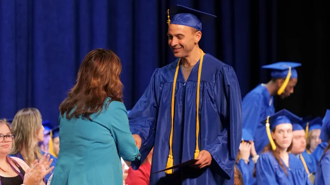 A person wearing a blue graduation cap and gown shakes hands with another individual in a teal suit on stage at a graduation ceremony. Several other people in similar attire are seated in the background.
