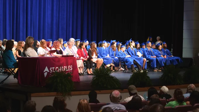 Graduates in blue caps and gowns sit on a stage next to a table with a St. Charles logo. An audience is seated in the foreground.