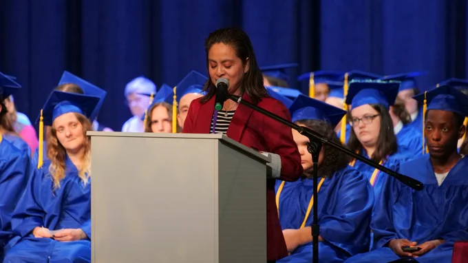 A person stands at a podium speaking into a microphone during a graduation ceremony. Participants wearing blue caps and gowns are seated on stage in the background.