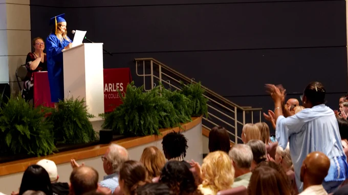 A graduate in a blue cap and gown stands at a podium on stage, smiling while holding a certificate. The audience, seated in an indoor auditorium, is applauding. A sign on the stage reads "St. Charles Community College".