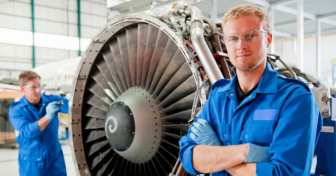 Two individuals in blue work uniforms are in an aircraft hangar. One stands confidently with arms crossed in front of a large jet engine, while the other works on an aircraft in the background. Both are wearing safety glasses and gloves.