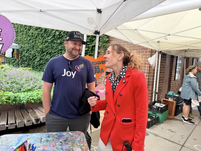 Elizabeth Shaffer speaks with a man wearing a Lake Irving hat at an Uptown market booth, with colorful community signs behind them.