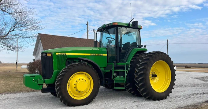 A John Deere tractor parked on a gravel driveway, with a house and trees in the background under a partly cloudy sky.