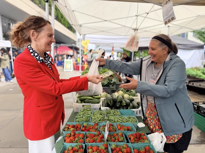 Elizabeth Shaffer buys fresh produce from a vendor at the Uptown Farmers Market, exchanging cash while holding a bag of vegetables.