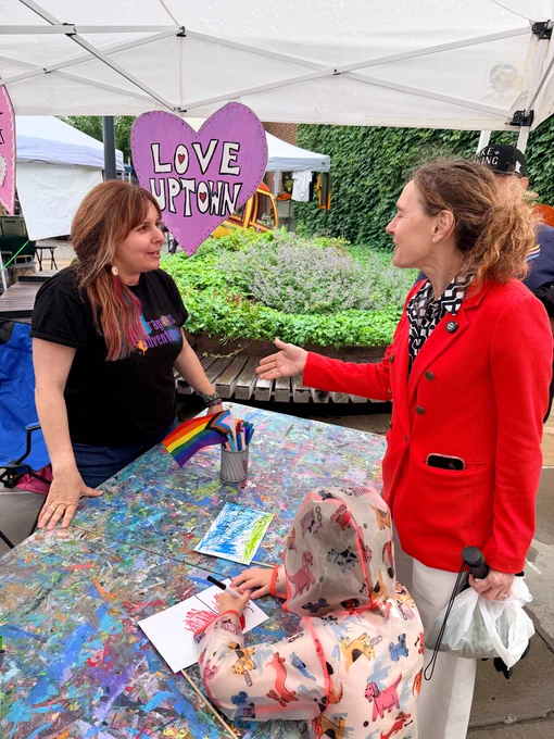 Elizabeth Shaffer talks with a vendor at an art table under a canopy, with a child drawing nearby and a “Love Uptown” sign in the background.