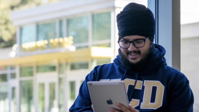 A male student wearing a UC Davis sweatshirt stands by a window in the daytime, looking at his e-reader and presumably reading digitally delivered course materials.