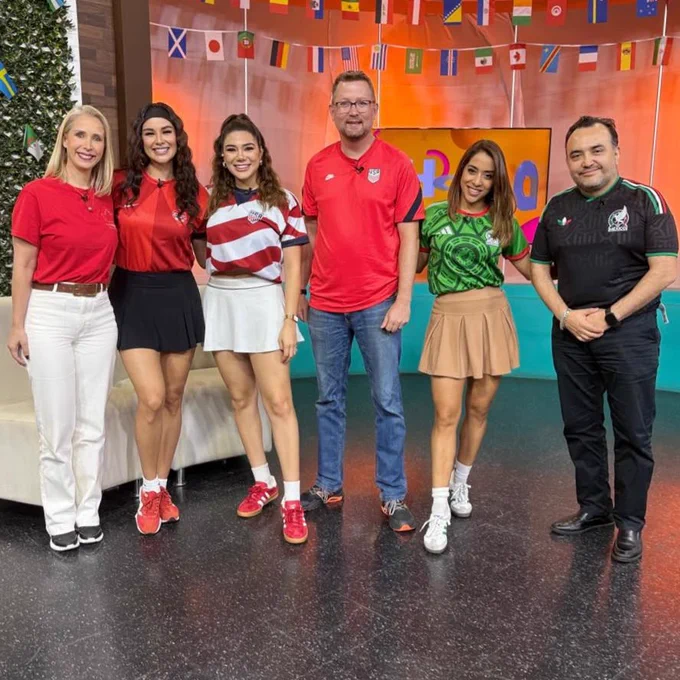 Six people stand in a brightly lit TV studio decorated with international flags. The group wears soccer jerseys and casual outfits in red, green, and black, posing together in front of a colorful set.