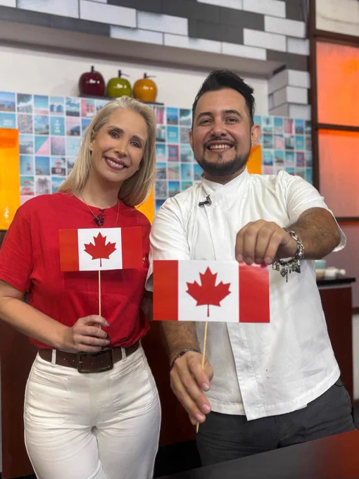 Two people in a TV studio hold small Canadian flags toward the camera. One person wears a red shirt and white pants, while the other wears a white chef-style shirt. A tiled wall and colorful décor appear in the background.