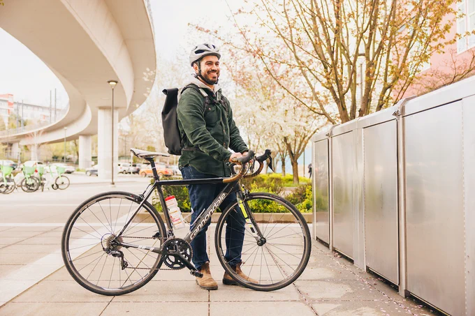 A person stands on a sidewalk next to a black bicycle and a row of silver bike lockers, wearing a white helmet and a dark jacket with a backpack. Blossoming trees and elevated train tracks are visible in the background.